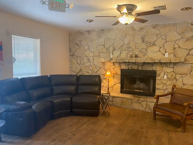 A cozy living room with a stone fireplace, a ceiling fan with a light, a black leather sectional sofa, a wooden armchair with brown cushions, a small side table with a lamp, and a window with blinds letting in natural light.