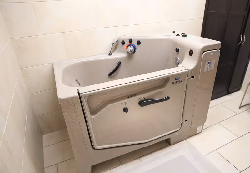 A beige walk-in bathtub with a side door and multiple control knobs, installed in a tiled bathroom with a dark wooden cabinet in the background.