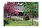 A single-story brick building partially obscured by green and purple-leaved trees, with a well-maintained lawn and shrubs in front.