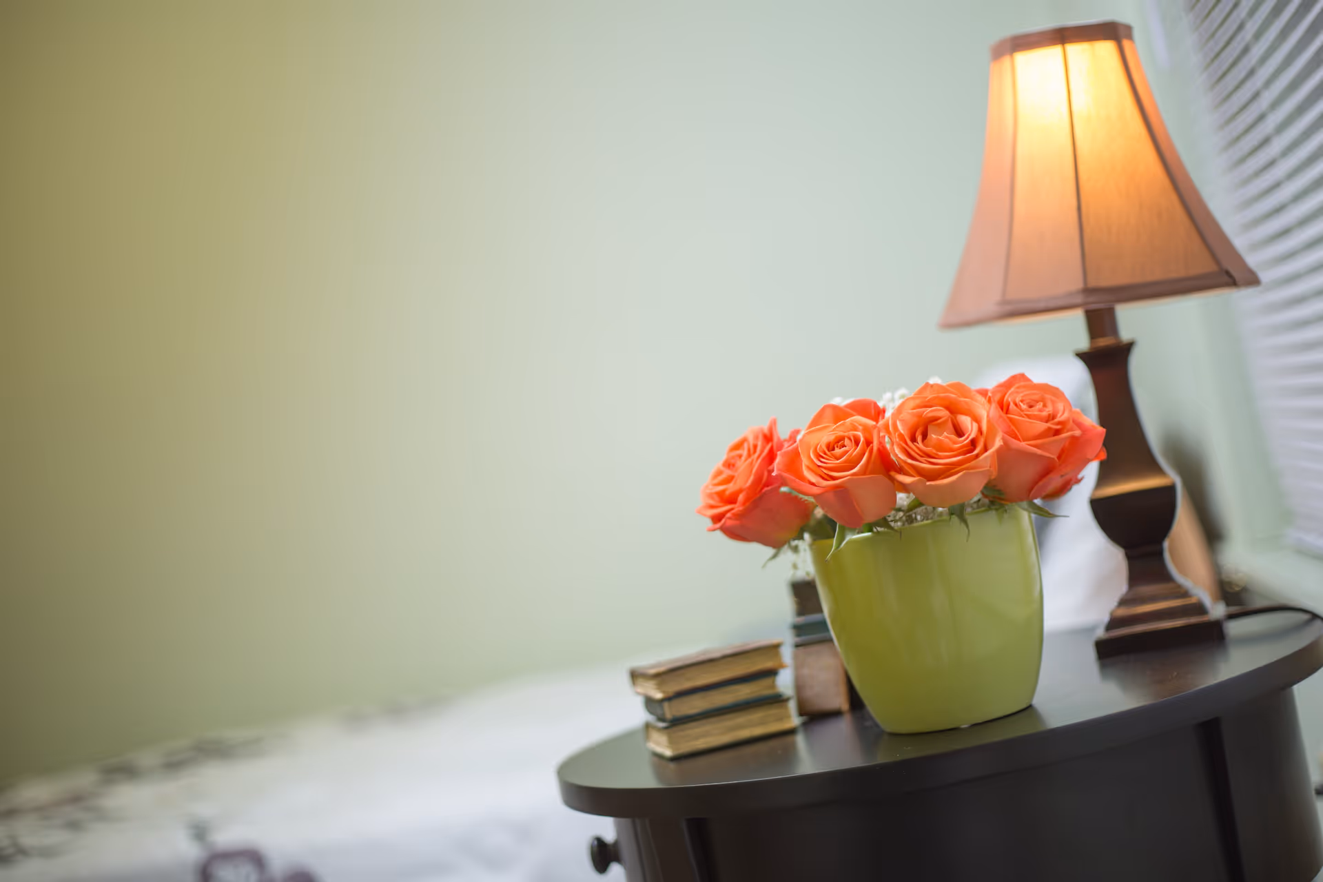 A close-up of a bedside table with a green vase holding orange roses, a stack of three old books, and a lit table lamp with a beige shade. In the background, part of a bed with white bedding and a light green wall are visible.