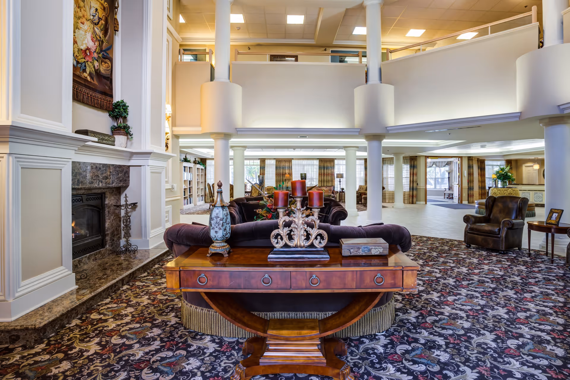 Spacious and elegant senior living facility common area with a patterned carpet, a wooden table with decorative items including a candelabra and a vase, plush sofas, a fireplace with a floral tapestry above it, and large windows with curtains allowing natural light to fill the room.