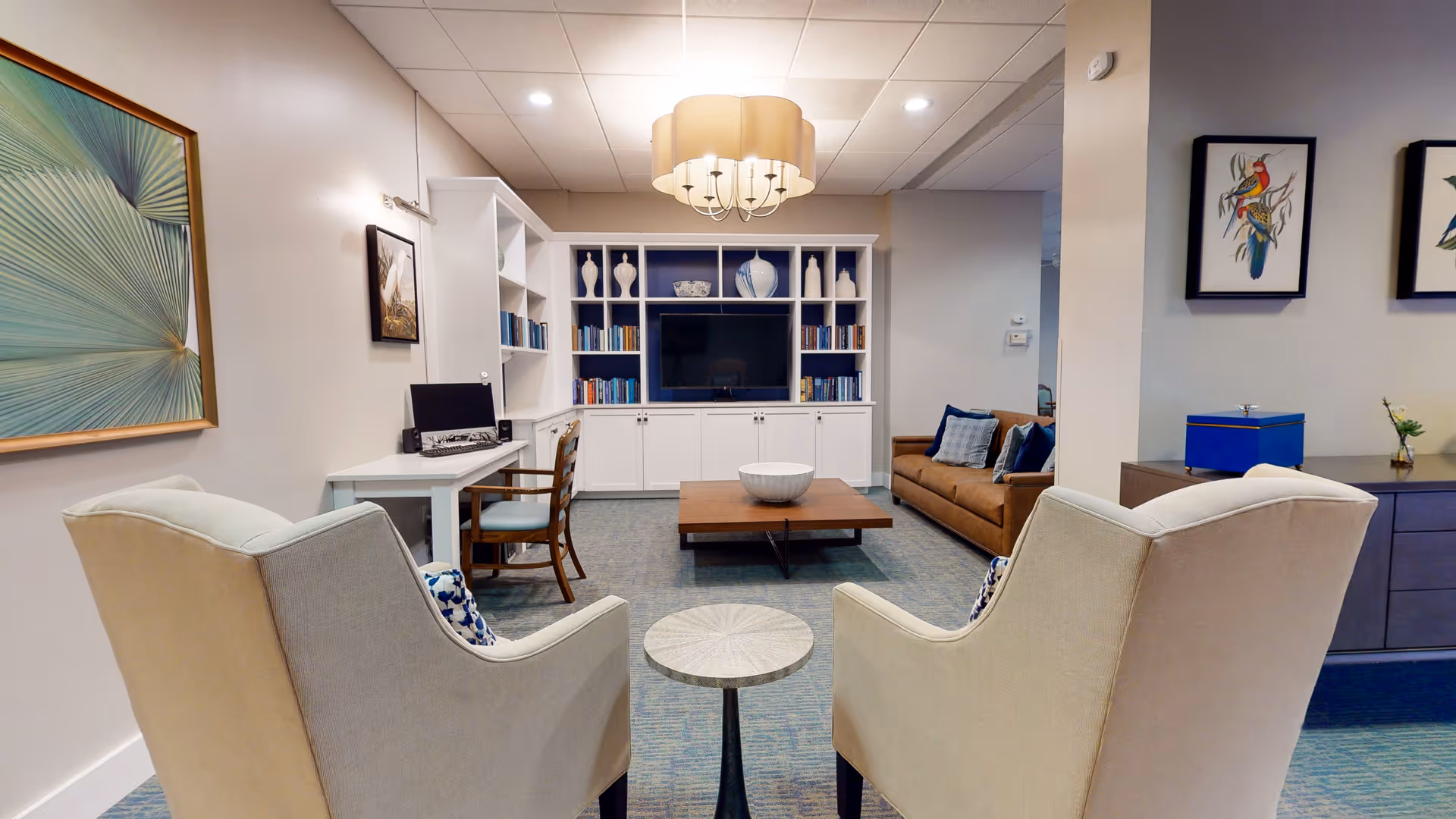 A cozy living room area with two beige armchairs facing a wooden coffee table and a built-in white shelving unit with a TV and decorative vases. There is a brown couch with blue pillows to the right, a small round side table between the armchairs, a desk with a computer and chair on the left, and framed artwork on the walls. The room is softly lit with a ceiling light fixture.