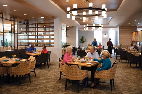 Seniors and staff conversing and eating at round tables in a bright, modern communal dining room with patterned chairs and overhead chandeliers.
