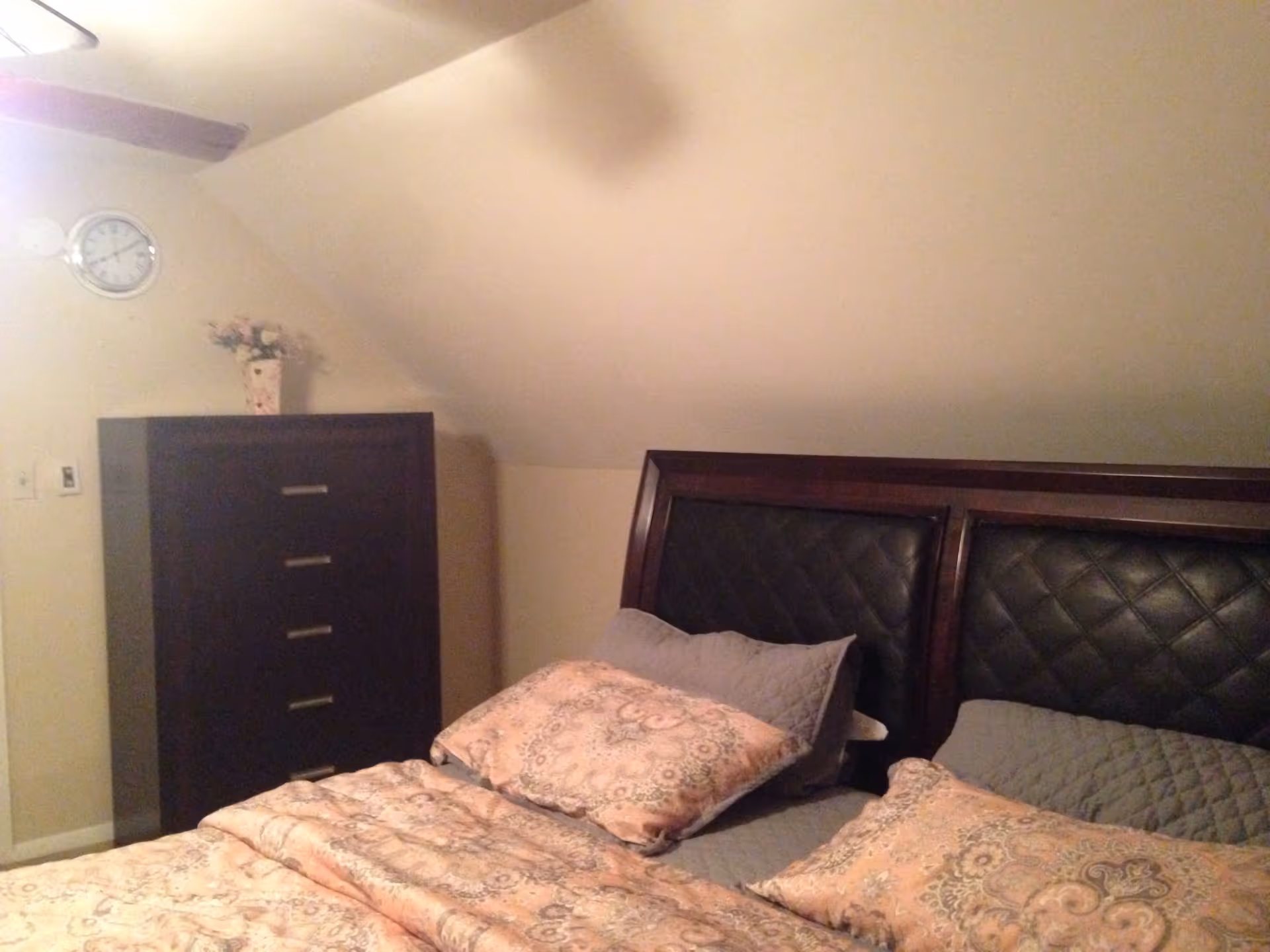 A bedroom with a bed featuring a dark wooden headboard with black padded panels, covered with a patterned quilt and pillows in shades of beige and gray. To the left of the bed is a tall dark wooden dresser with five drawers, topped with a small floral arrangement in a vase. A round wall clock is mounted on the wall above the dresser. The ceiling slopes downward toward the dresser side of the room.