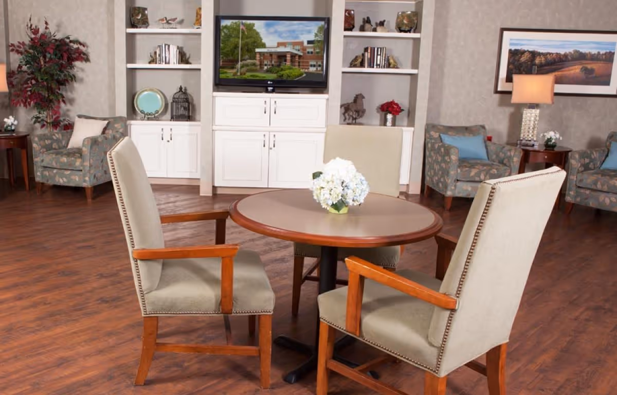 A cozy sitting area with a round wooden table surrounded by four upholstered chairs with wooden arms. In the background, there are built-in white shelves and cabinets, a flat-screen TV displaying an image of a building, two armchairs with floral upholstery and blue cushions, a side table with a lamp, and a framed landscape picture on the wall. The floor is wooden, and there is a potted plant in the corner.