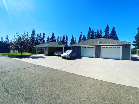 Single-story light-gray care home with two garage doors, parked cars and a wide concrete driveway under a bright blue sky.