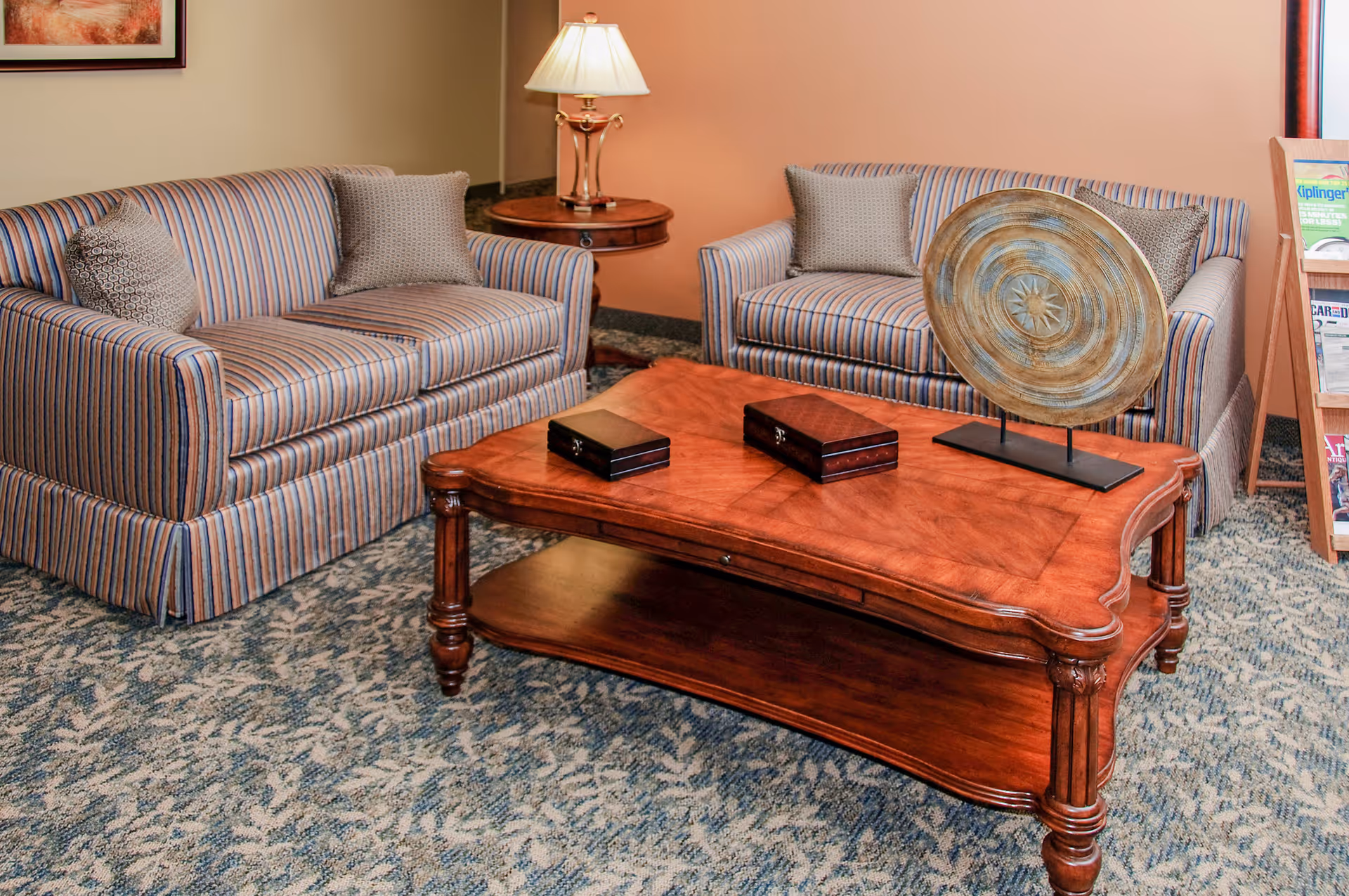 A cozy living room area with two striped sofas facing each other, each with patterned throw pillows. Between them is a wooden coffee table with decorative boxes and a large ornamental plate. A round side table with a lamp is positioned behind one sofa, and a magazine rack is visible in the corner.