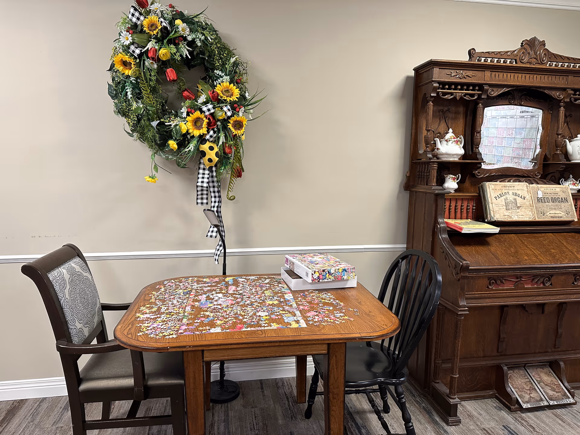 A wooden table with two chairs, one upholstered and one black wooden, with a partially completed jigsaw puzzle and puzzle box on top. Behind the table is a decorative floral wreath with sunflowers and red flowers hanging on a beige wall. To the right is an ornate wooden cabinet with vintage sheet music and ceramic teapots displayed.