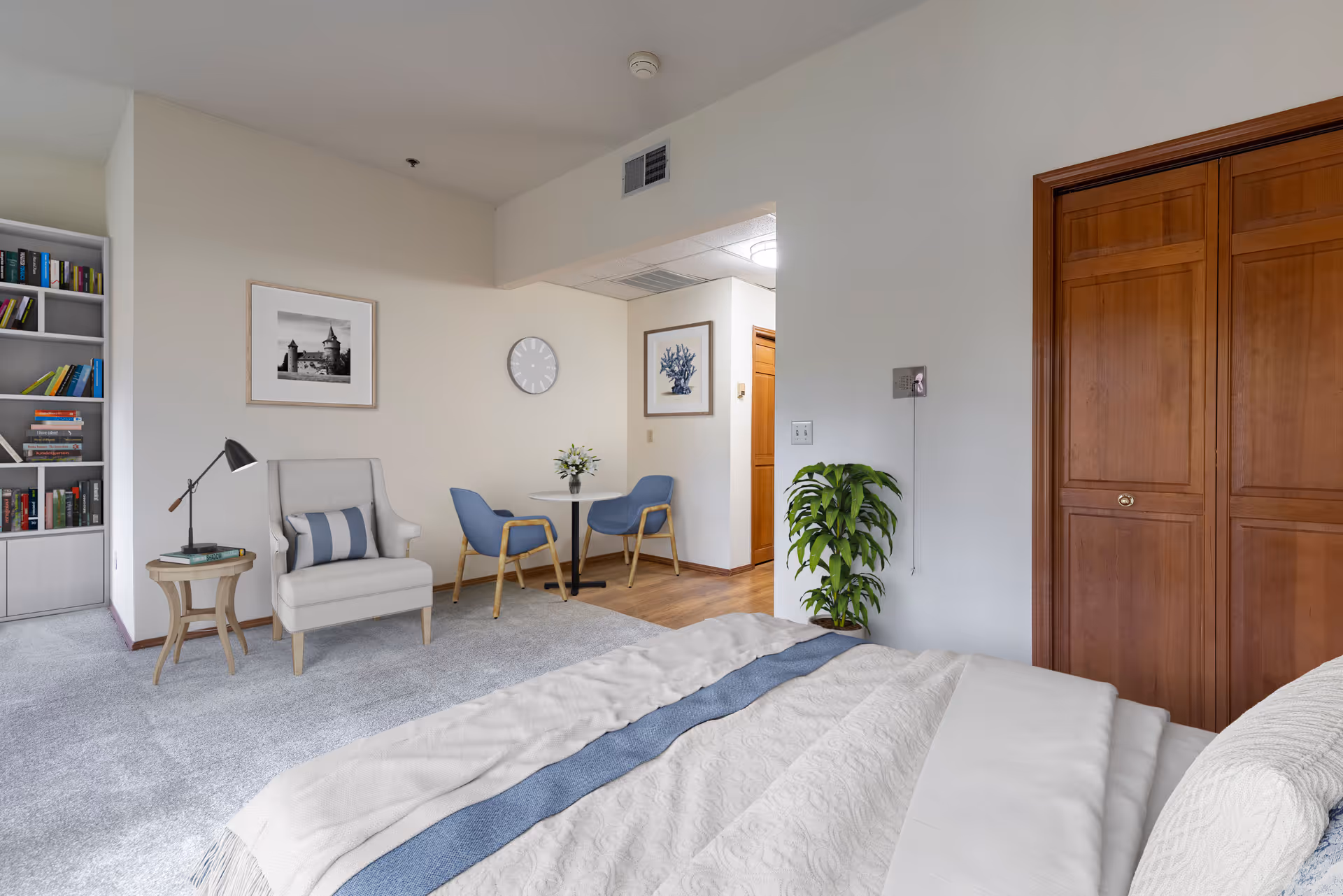 Bright furnished bedroom with a bed in the foreground, an armchair and side table, a small dining table with chairs, bookshelf, and wooden closet doors.