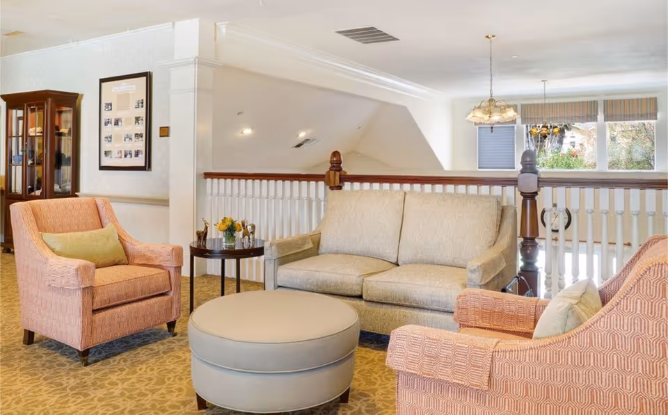 A cozy senior living facility lounge area with a beige sofa, two patterned armchairs with green cushions, a round ottoman, and a small side table with flowers and decorative items. The room has carpeted flooring, white walls, a wooden railing, and large windows letting in natural light.