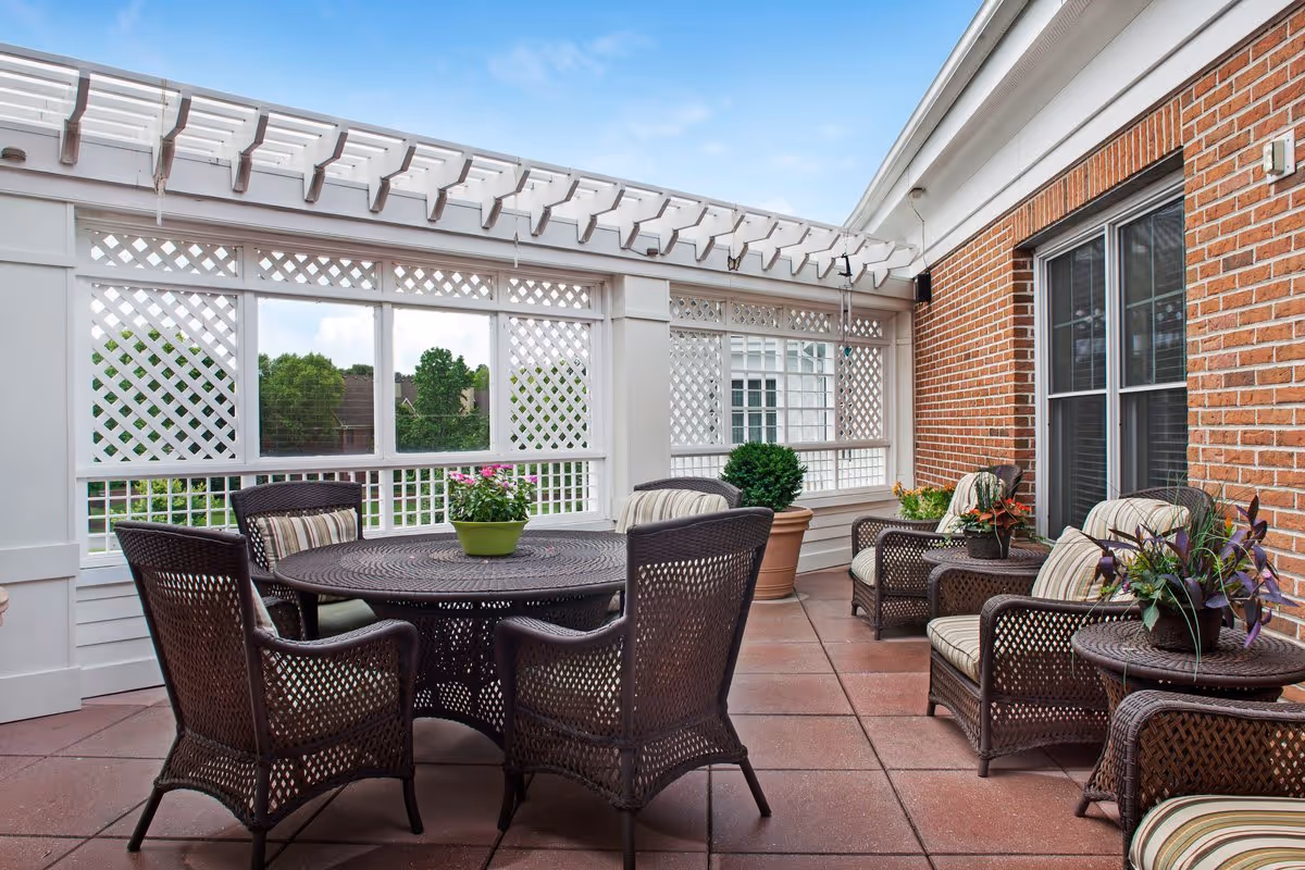 Outdoor patio area with brown wicker furniture including a round table with four chairs and three cushioned armchairs with side tables. The patio has a white lattice fence and a pergola overhead, with potted plants and a brick wall with windows on one side.