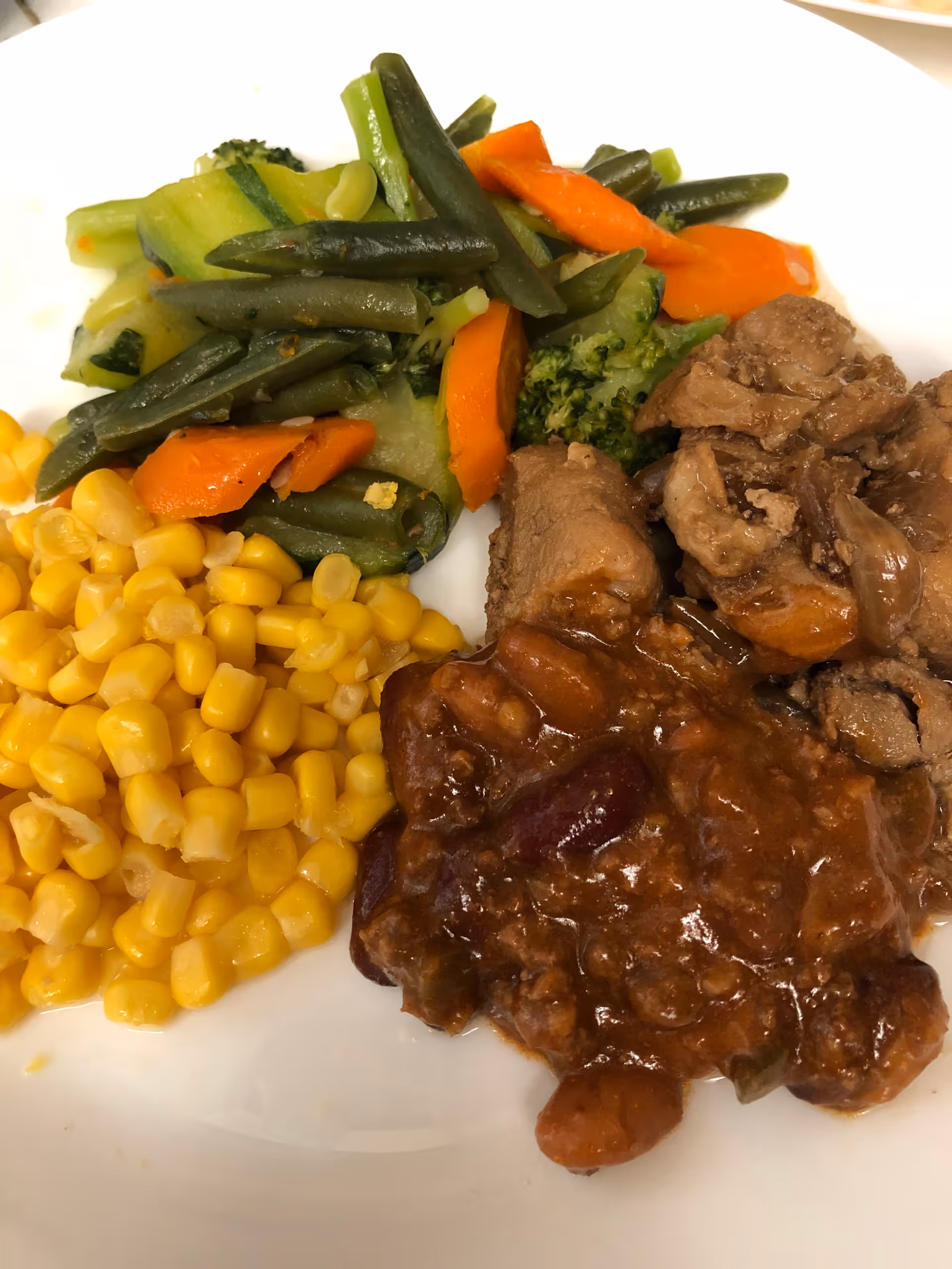 A plate with corn, mixed steamed vegetables, chunks of meat and a saucy bean or chili side.