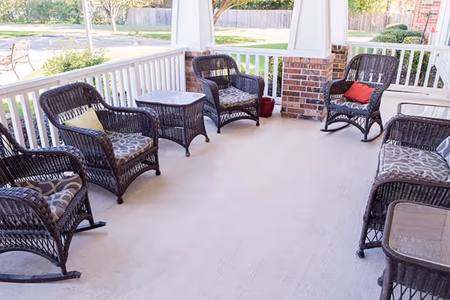 A covered outdoor patio area with six dark wicker chairs arranged in a semi-circle, each with patterned cushions and some with colorful throw pillows. There are two small wicker tables between the chairs. The patio overlooks a grassy area with trees and a parking lot in the background.