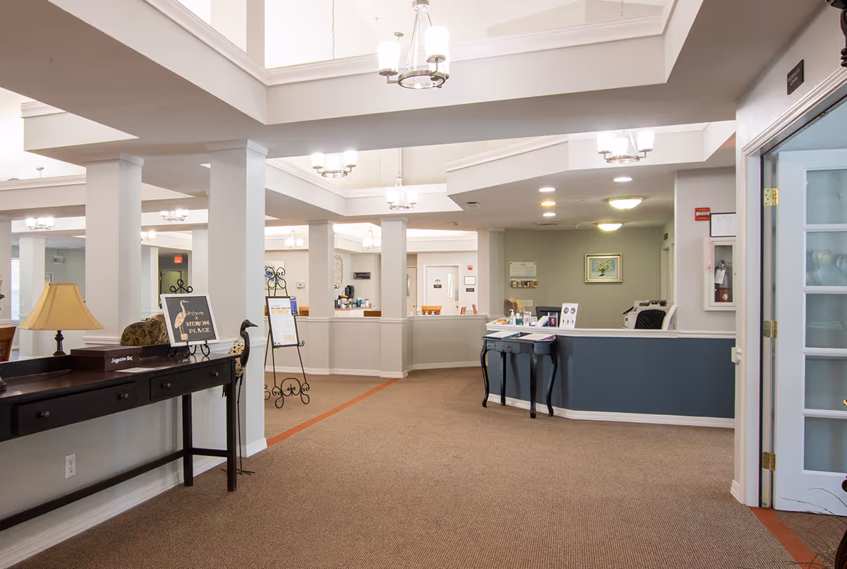 Spacious senior living facility reception area with a welcoming front desk, decorative tables, columns, and ceiling lights.