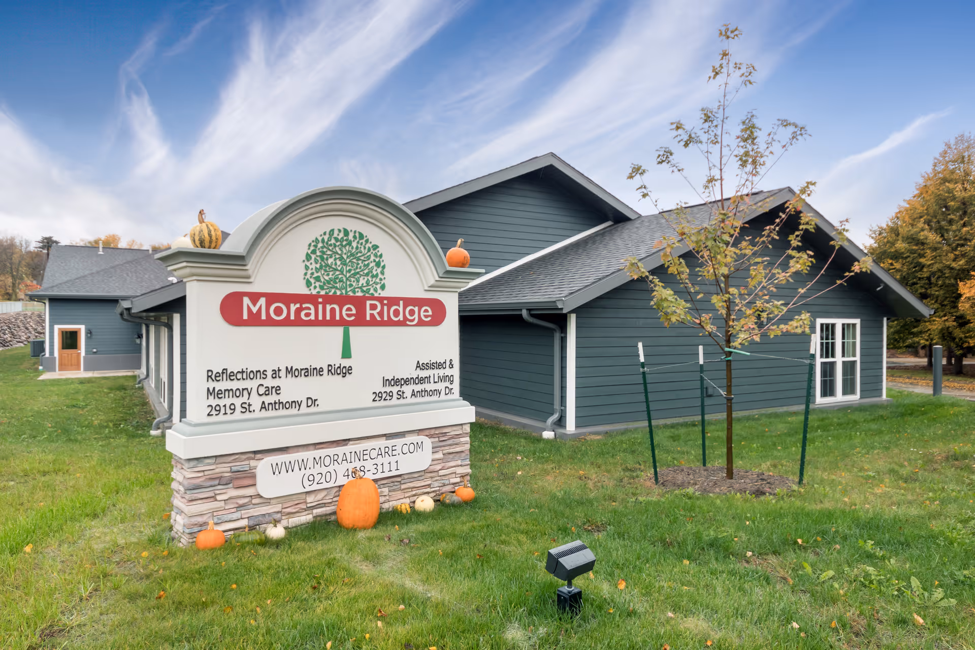 A large sign reading 'Moraine Ridge' and 'Reflections at Moraine Ridge Memory Care' sits on the lawn in front of a gray single-story building with pumpkins and a small tree.