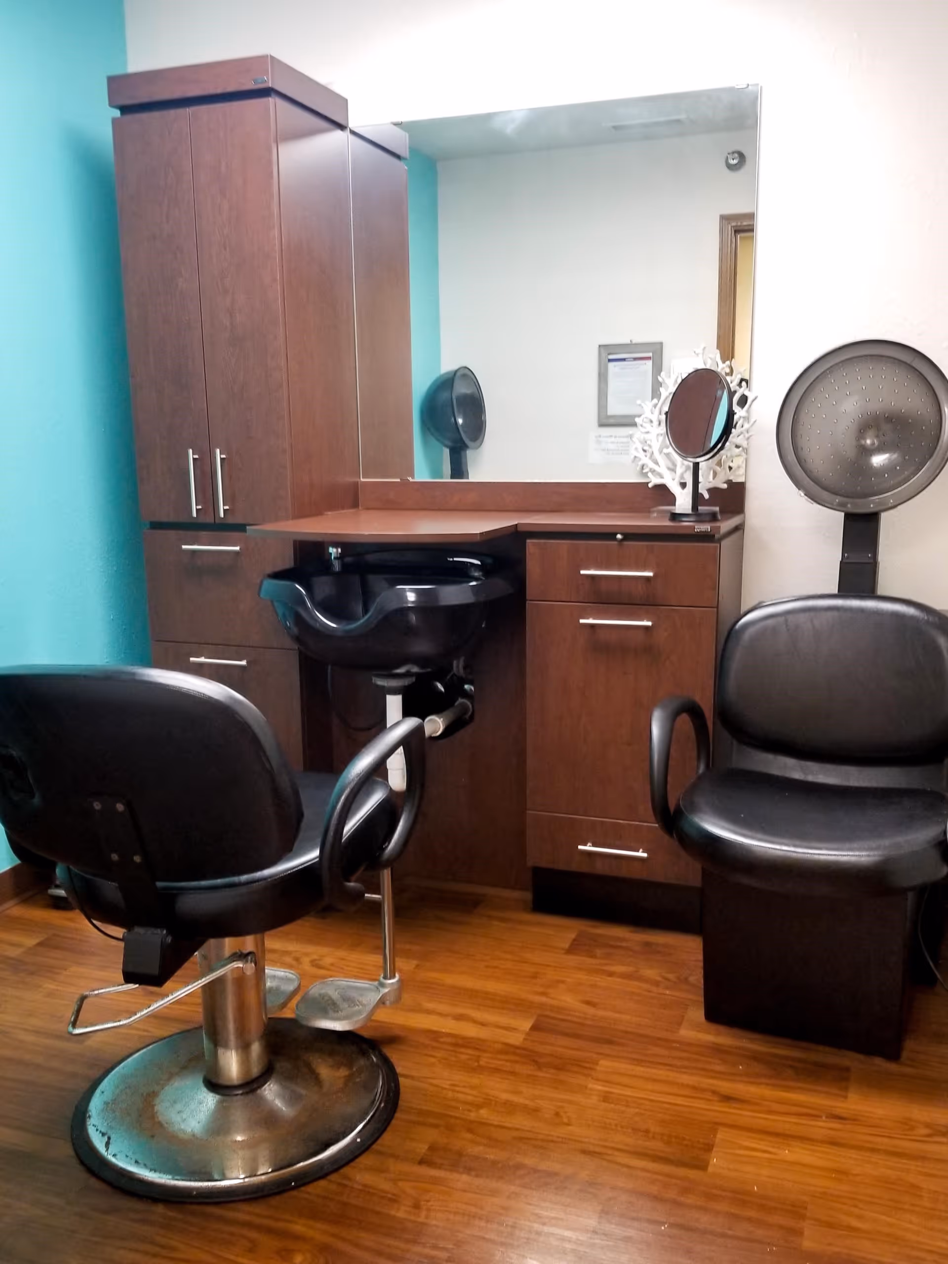 Interior view of a salon area with a black salon chair in front of a black hair washing sink, a wooden cabinet with drawers and a large mirror above it. To the right, there is a black hair dryer chair with a hooded hair dryer attached. The floor is wooden and the walls are painted teal and white.