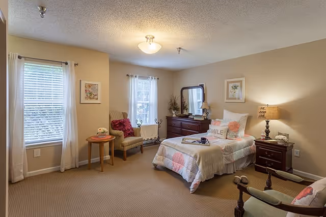 A cozy bedroom with a single bed covered in a quilted bedspread with pink and white patterns. The bed has pillows, including one with the word 'SMILE'. There is a wooden nightstand with a lamp and decorative items beside the bed. A wooden dresser with a mirror and a lamp is against the wall. Two windows with white curtains allow natural light into the room. A cushioned armchair with a red pillow and a small round wooden table with a flower vase are near one window. The walls are painted beige and the floor is carpeted.