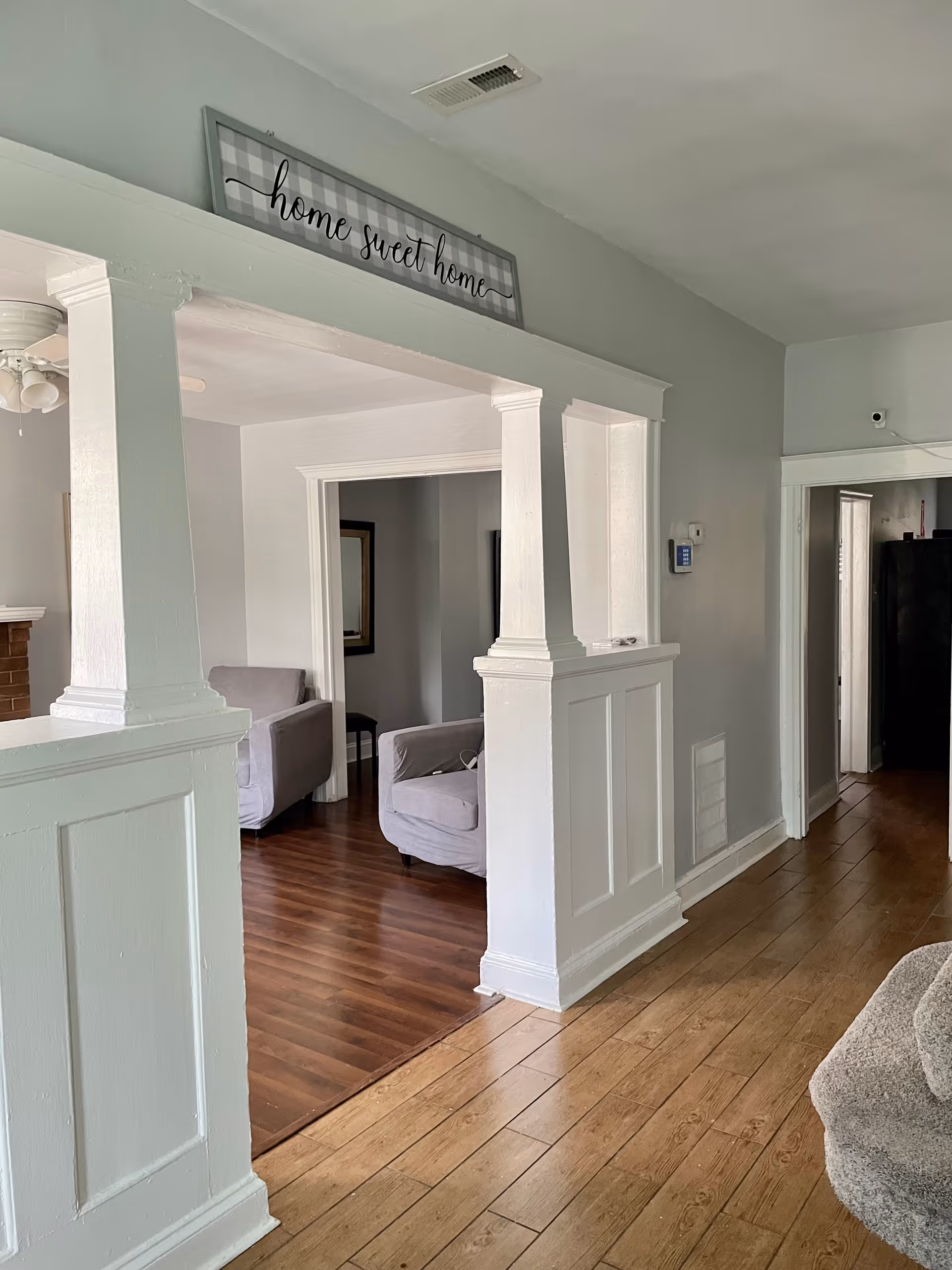 Open interior view of a living room and hallway with white columns, wood floors, and a 'home sweet home' sign.