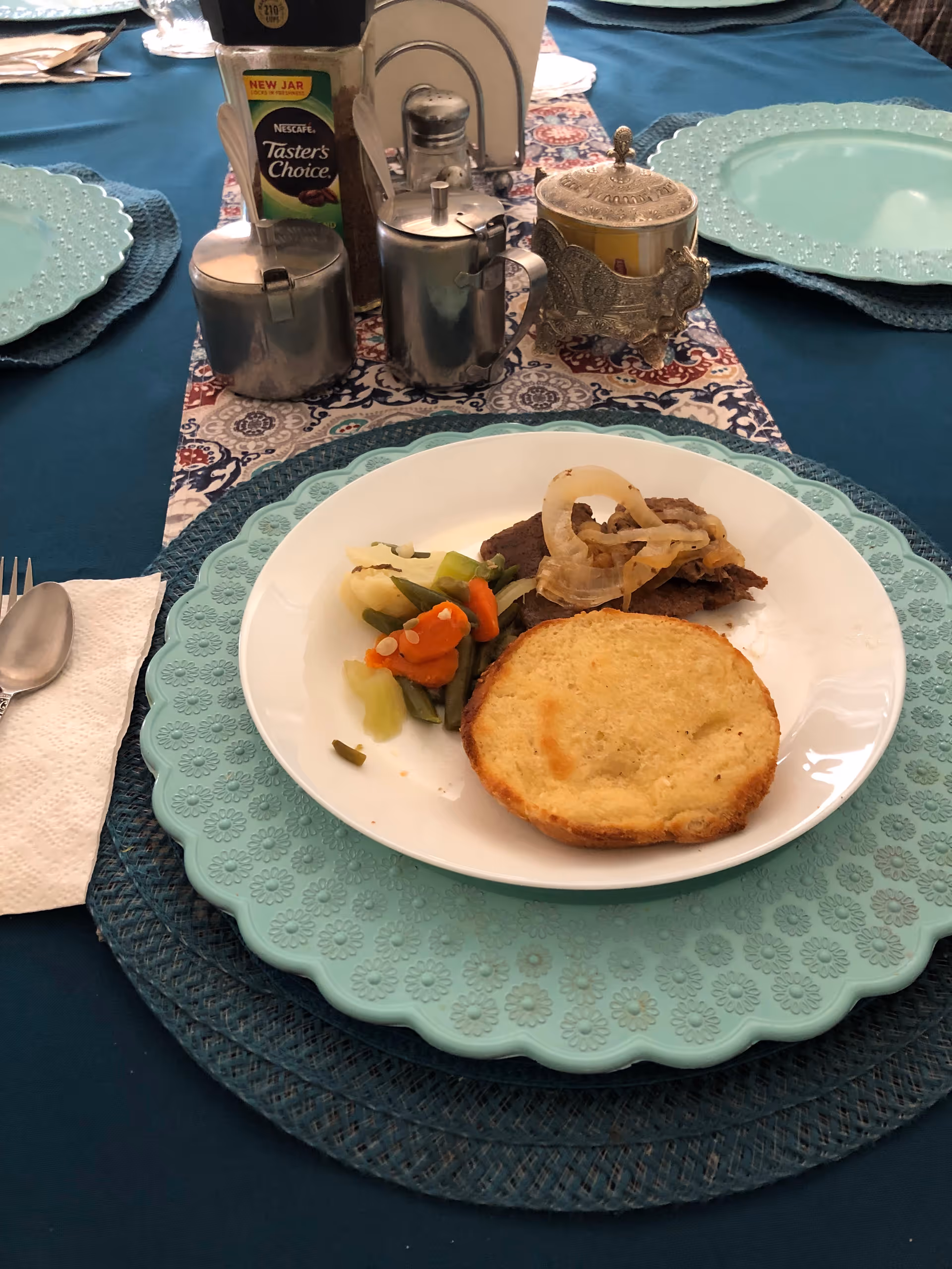 A white plate with a serving of cooked vegetables, a piece of meat topped with sautéed onions, and a toasted bread slice on a blue floral-patterned placemat. The table is covered with a blue tablecloth and has silver condiment containers, a salt shaker, and a decorative container in the background.