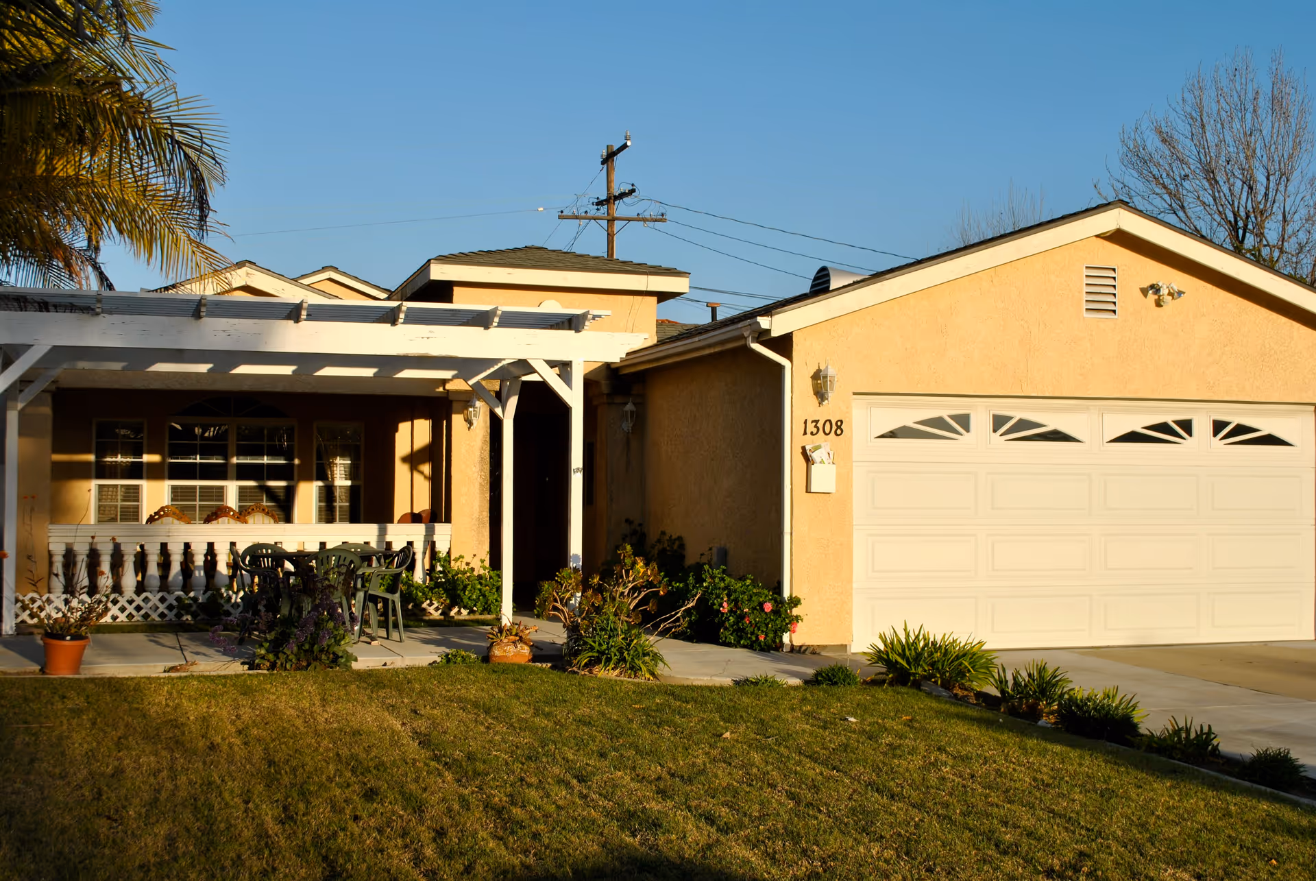 Exterior view of a single-story residential building with a beige stucco finish, a white garage door, and a covered front porch with outdoor chairs and a table. The house number 1308 is visible near the garage. There are plants and a well-maintained lawn in the front yard under a clear blue sky.