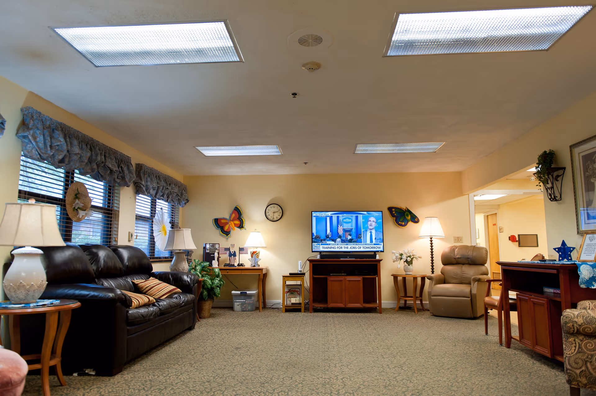 A cozy living room area in a senior living facility with a black leather sofa, a beige recliner chair, multiple lamps, a television mounted on a wooden cabinet, butterfly wall decorations, and windows with blue valances.