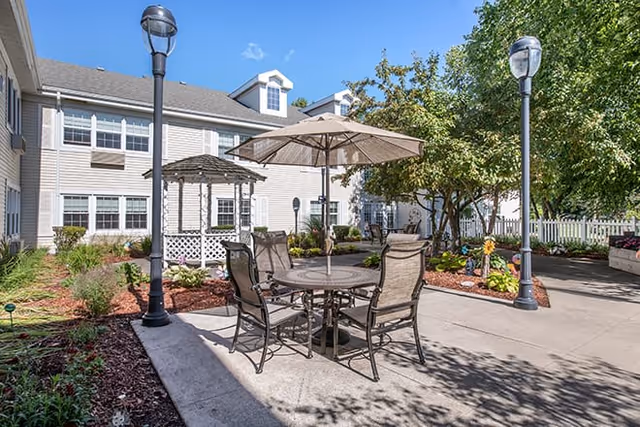 Outdoor patio area at Brookdale Utica Assisted Living with a round table, four chairs, and a large umbrella. The area is surrounded by landscaped gardens, trees, and a white gazebo. Two tall lamp posts are visible, and the building with multiple windows is in the background under a clear blue sky.