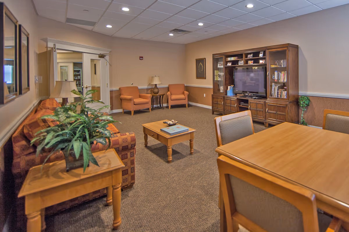 A cozy living room area in a senior living facility featuring a patterned sofa, two orange armchairs, a wooden coffee table with books, a wooden entertainment center with a TV and shelves filled with books and photos, a wooden dining table with chairs, and a potted plant on a side table. The room has beige walls, carpeted floor, and recessed ceiling lights.