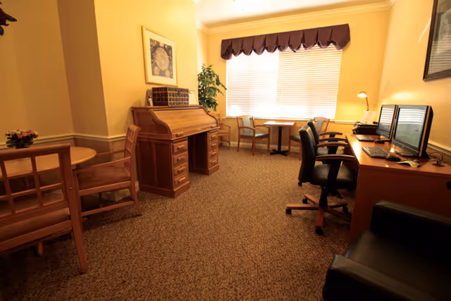 A warm, carpeted common room with a roll-top desk, computer workstations, tables and chairs beneath a large window.