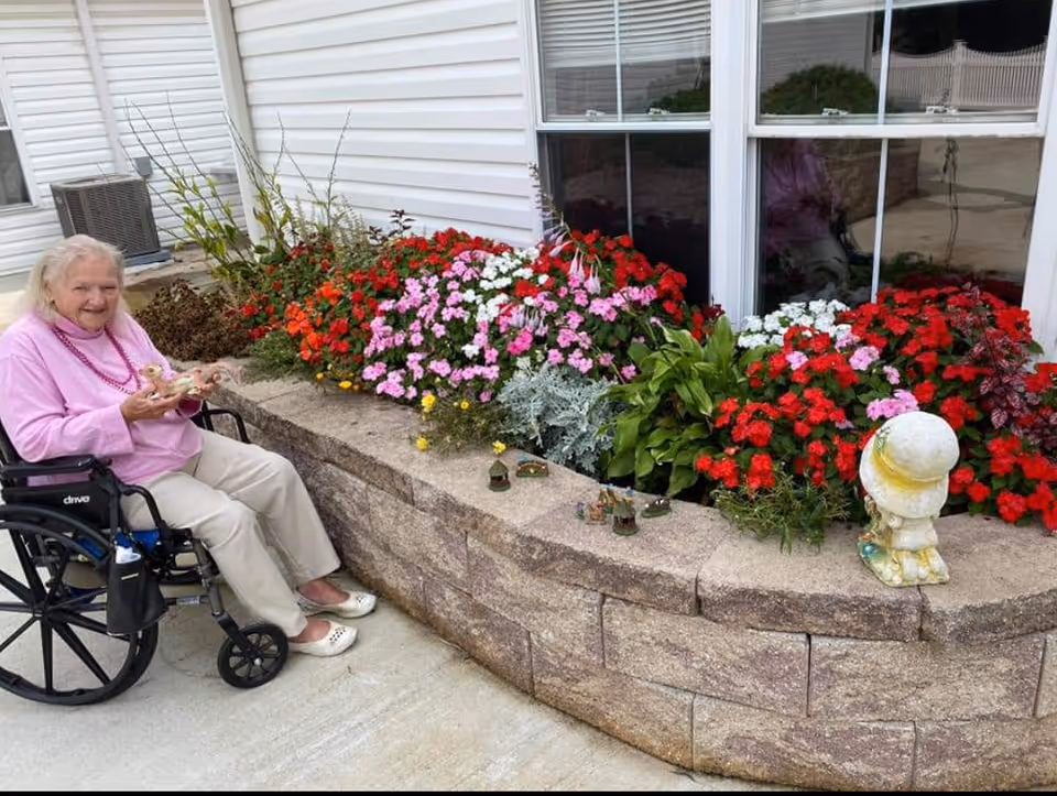 An elderly woman in a wheelchair wearing a pink sweater and beige pants is sitting outside next to a raised flower bed filled with colorful red, pink, white, and green plants. The flower bed is made of stone blocks and is situated against the exterior wall of a building with white siding and windows. Small decorative figurines are placed on the edge of the flower bed.