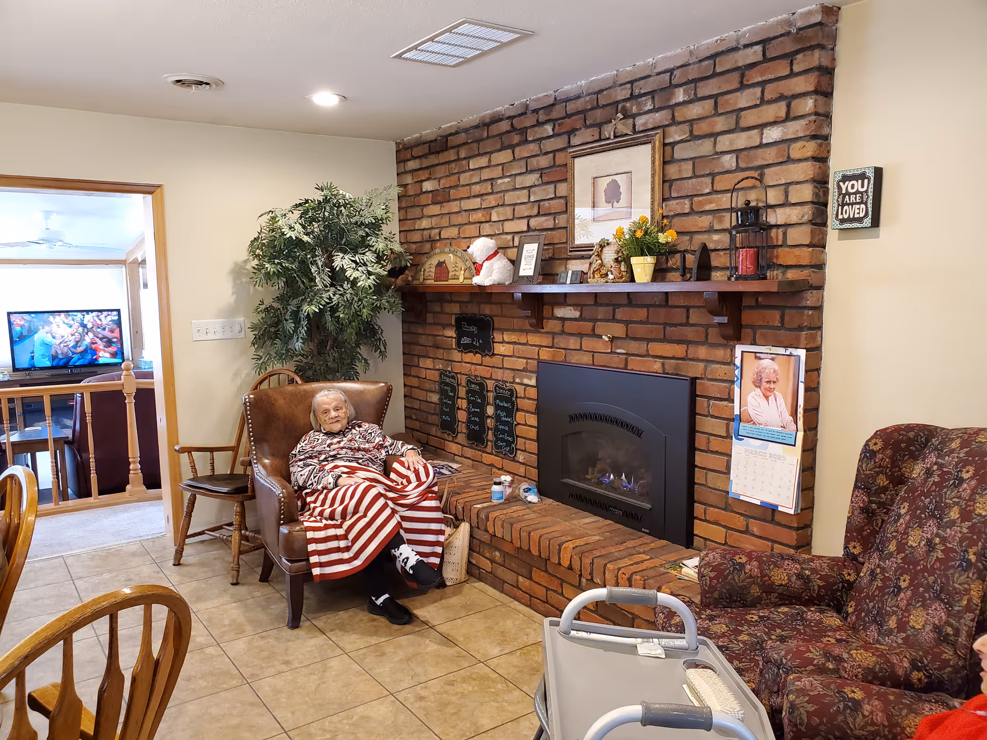 An elderly resident sits wrapped in a red-and-white striped blanket in a cozy living room with a brick fireplace, armchairs, and a walker.