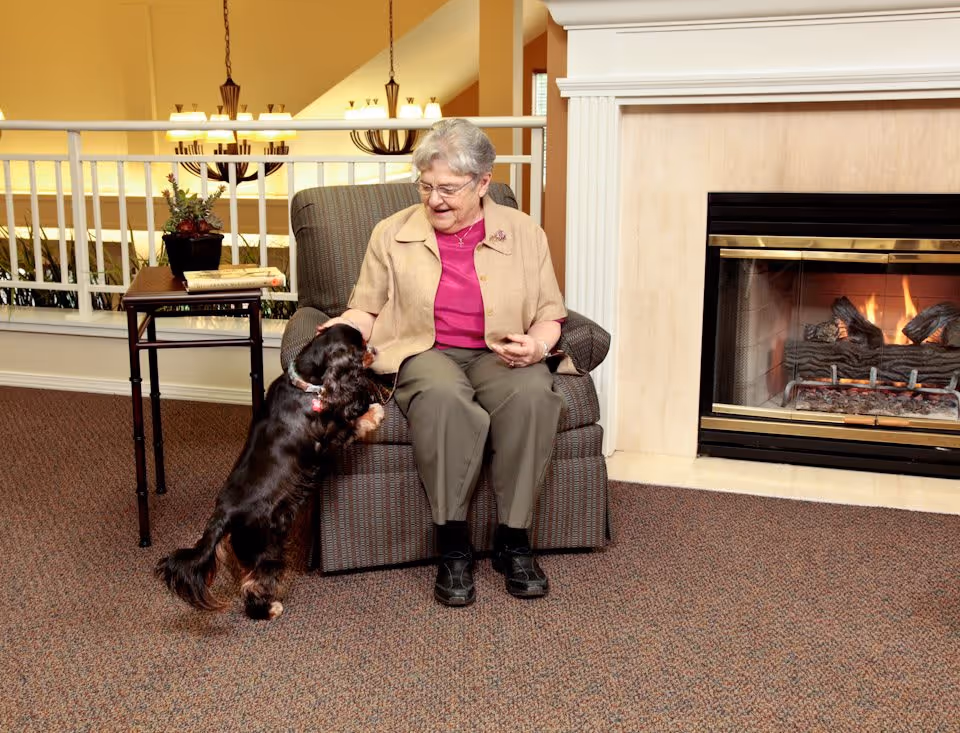An elderly woman sitting in a cushioned armchair petting a small black and brown dog standing on its hind legs in front of her. They are in a cozy room with a lit fireplace to the right and a small table with a plant and books behind the chair. Warm lighting and carpeted floor create a comfortable atmosphere.