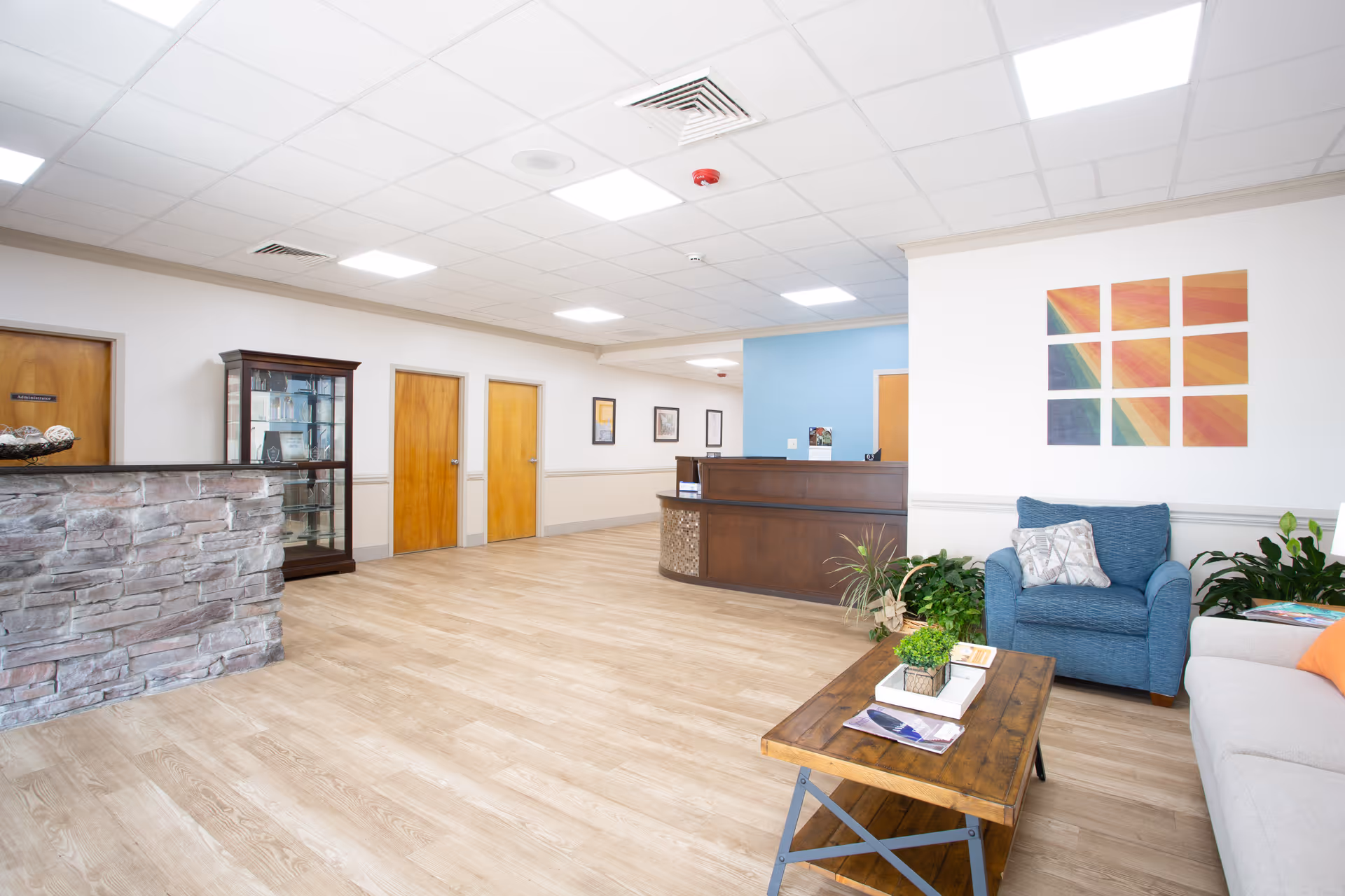 A bright and spacious reception area with light wood flooring, a stone-faced reception desk on the left, a wooden reception counter straight ahead, and a seating area on the right featuring a blue armchair, a beige sofa, and a wooden coffee table with plants and magazines. The walls are white with colorful abstract artwork and framed pictures, and there are several wooden doors along the back wall.
