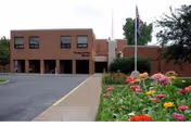 Exterior view of Presbyterian Manors III building with a paved driveway and a garden with colorful flowers in the foreground. An American flag is visible near the entrance.