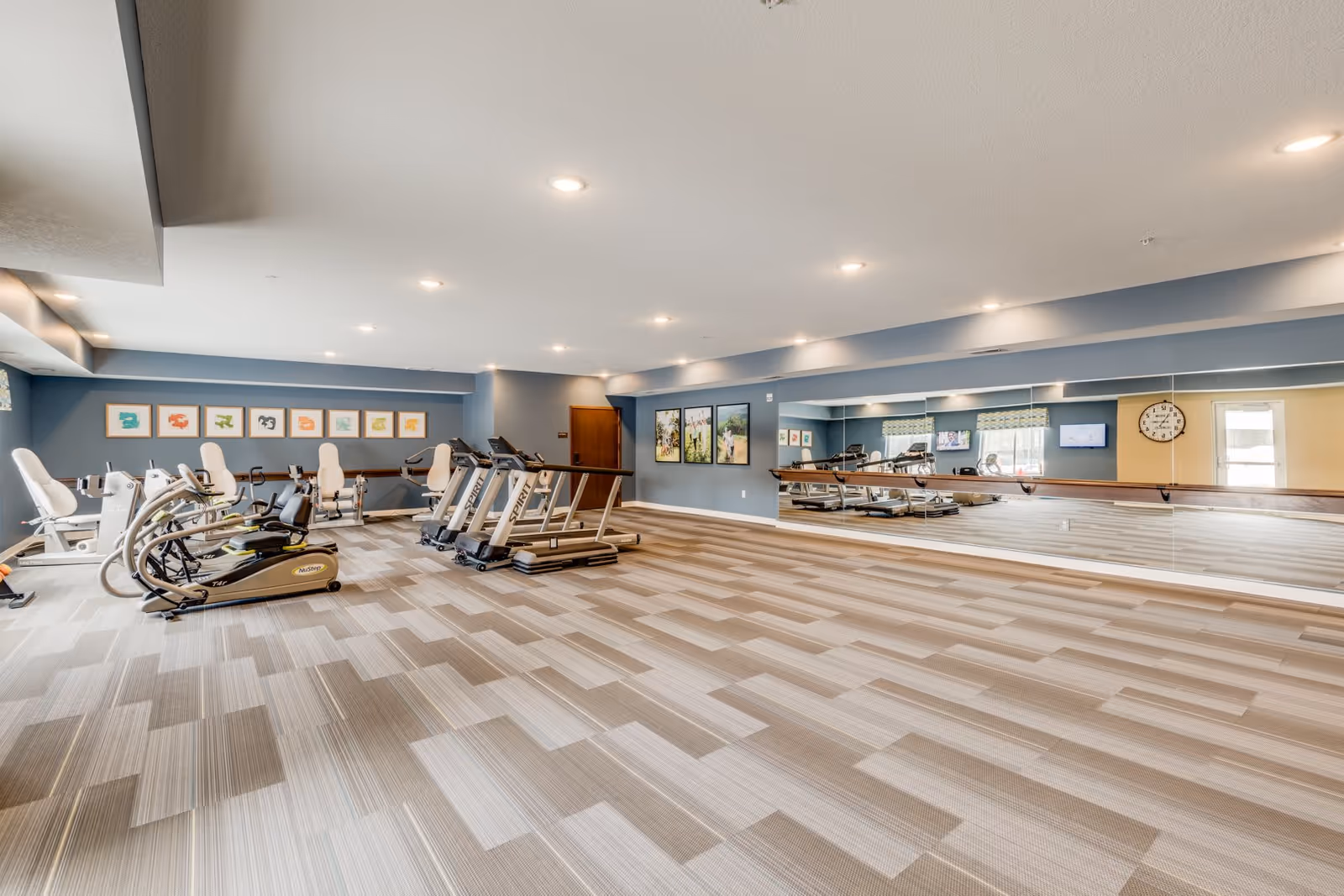 Spacious fitness room with treadmills, elliptical machines, a large wall mirror, and patterned carpet in a senior living facility.