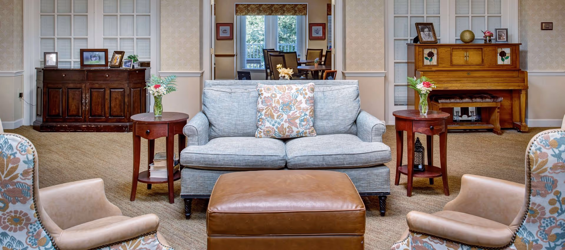 Bright, traditionally decorated common living room with a light-blue sofa, leather ottoman, patterned armchairs, side tables with flowers, and a piano against the wall.