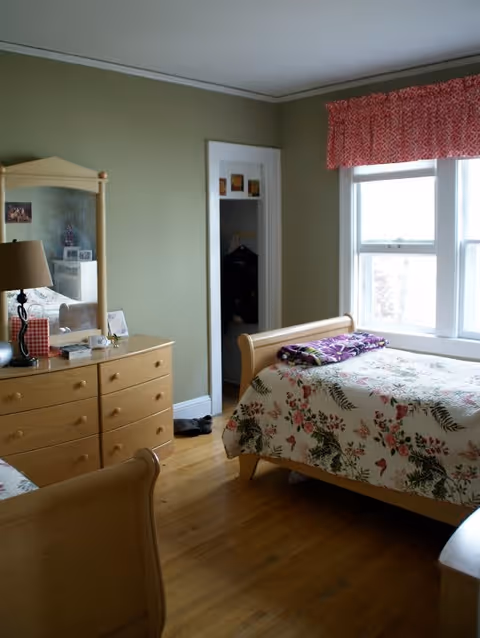 A bedroom with light green walls and wooden floor. There is a wooden bed with a floral bedspread and a folded blanket on top. Next to the bed is a wooden dresser with a mirror, a lamp, and some small items on it. A window with a red patterned valance lets in natural light. An open closet door is visible in the background.