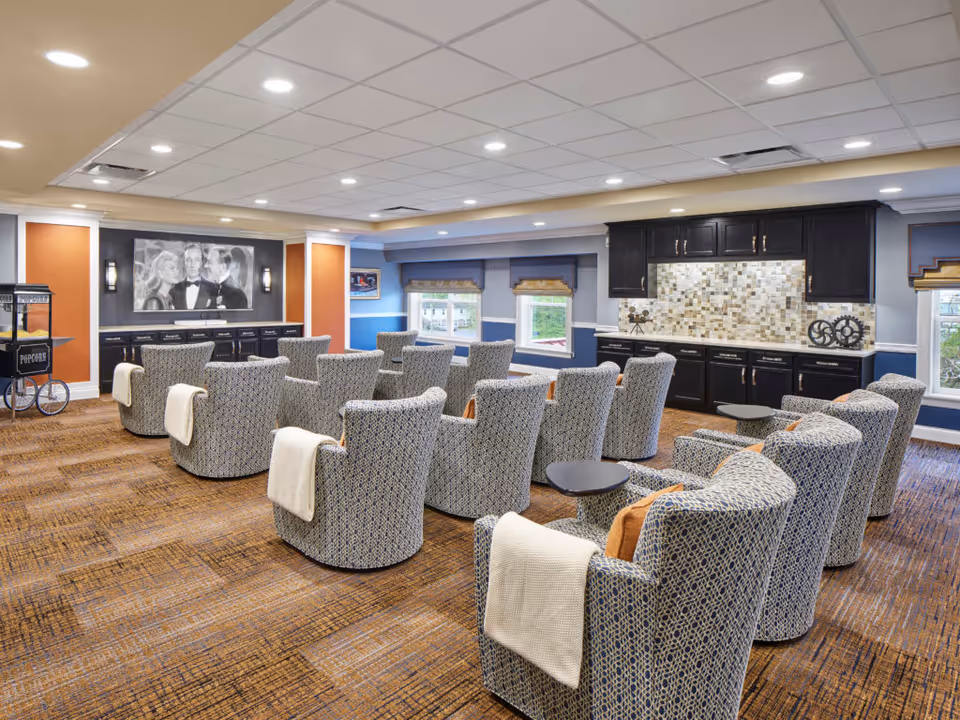 A cozy senior living facility common room with multiple patterned armchairs arranged in rows facing a large black and white movie poster on the wall. The room features a popcorn machine on the left, a kitchenette with dark cabinets and a mosaic tile backsplash on the right, and several windows letting in natural light.