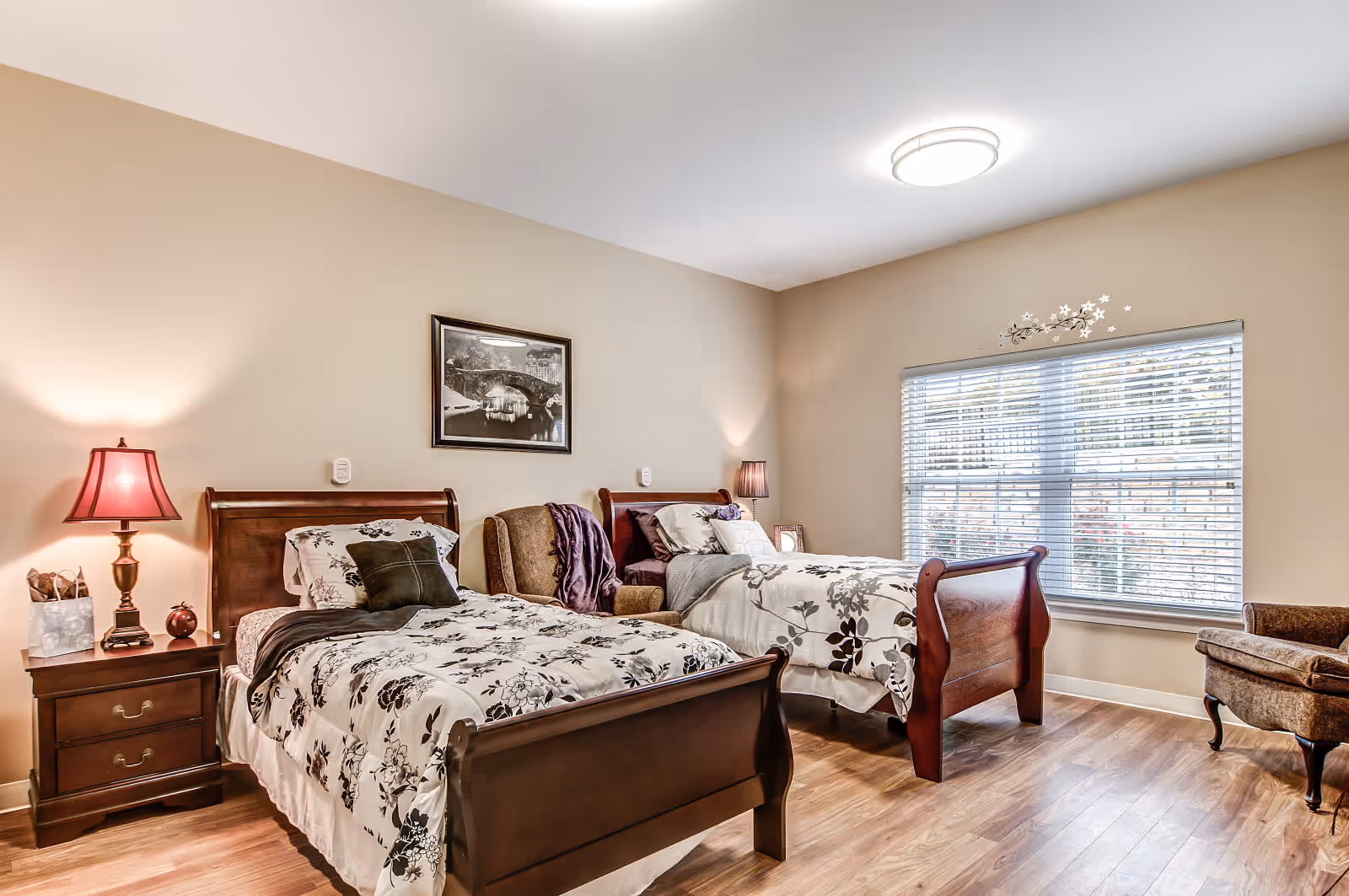 A bright bedroom with two wooden twin beds, each with floral patterned bedding. Between the beds is a brown upholstered armchair with a purple throw blanket. A wooden nightstand with a red lamp and decorative items is beside one bed. A large window with blinds lets in natural light, and a framed black and white picture hangs on the beige wall above the beds. The room has wood flooring and a modern ceiling light.