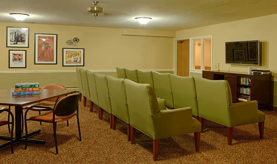 A cozy common room with two rows of green upholstered chairs facing a wall-mounted flat-screen TV. To the left, there is a wooden table with four chairs and several board games stacked on top. The walls are decorated with framed vintage movie posters and a popcorn sign. The room has warm lighting and carpeted flooring.