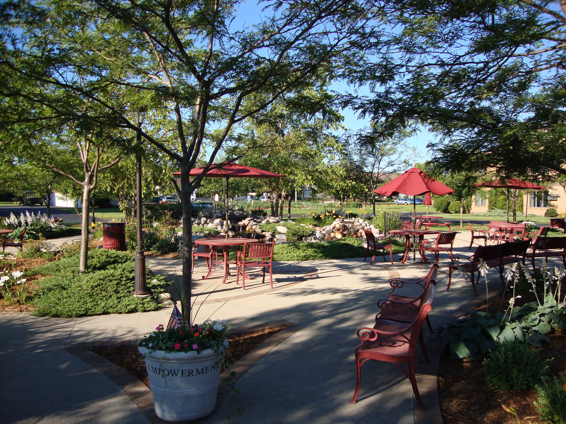 Sunny landscaped outdoor courtyard with red metal tables, chairs, umbrellas and benches among trees, planters and walking paths.