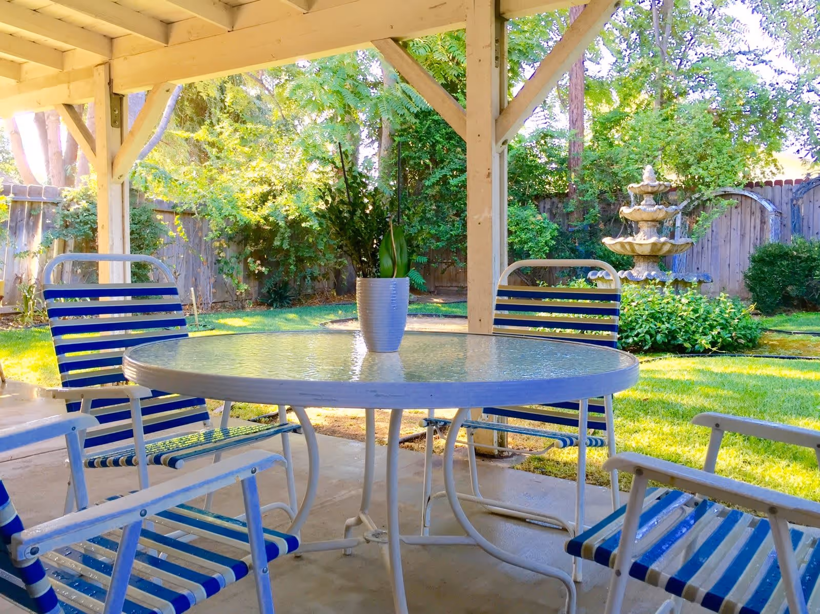 Outdoor covered patio area with a round glass table and four blue and white striped metal chairs. A small potted plant is placed in the center of the table. In the background, there is a garden with green grass, bushes, trees, a wooden fence, and a multi-tiered stone water fountain.