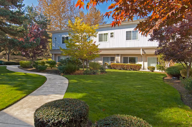 A well-maintained outdoor garden area with a curved concrete pathway leading through green grass, bushes, and trees with autumn-colored leaves in front of a two-story building with white siding and multiple windows.
