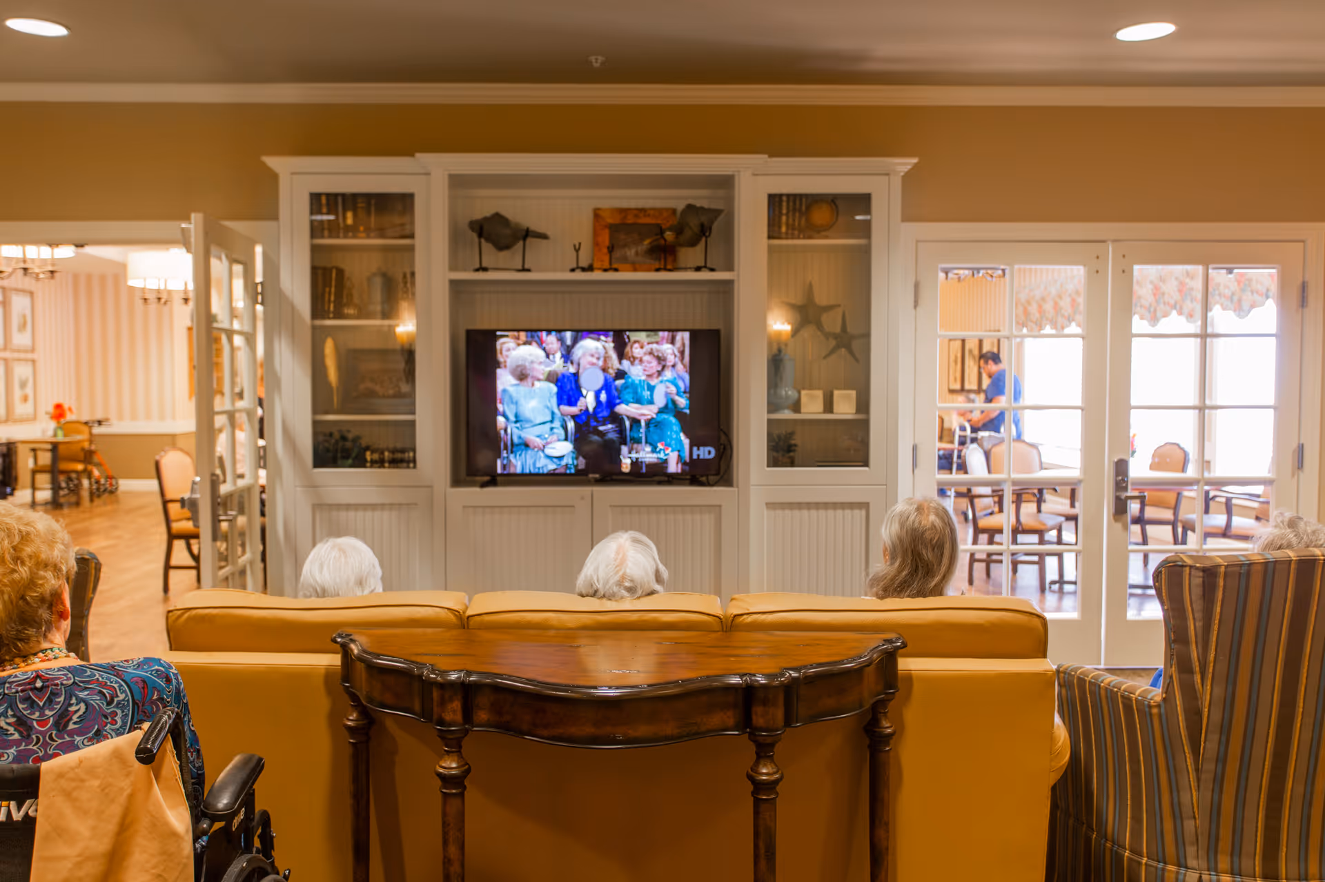 A group of elderly people sitting on couches and chairs in a living room area watching a television mounted in a white built-in cabinet. The room has warm lighting and a wooden table behind the couch. Through glass doors, another room with tables and chairs is visible, where a person is standing.
