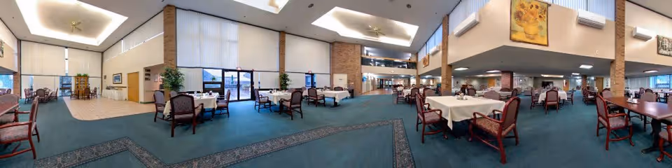 Spacious dining room with multiple tables covered with white tablecloths and surrounded by wooden chairs with cushioned seats. The room has high ceilings with recessed lighting, large windows allowing natural light, and decorative plants. The carpet is green with patterned borders, and there are brick columns and framed artwork on the walls.