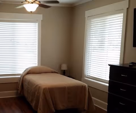 A small bedroom with a single bed covered in a beige blanket, positioned between two windows with white blinds. There is a ceiling fan with a light fixture above, a small bedside table with a lamp, and a dark wooden dresser on the right side.