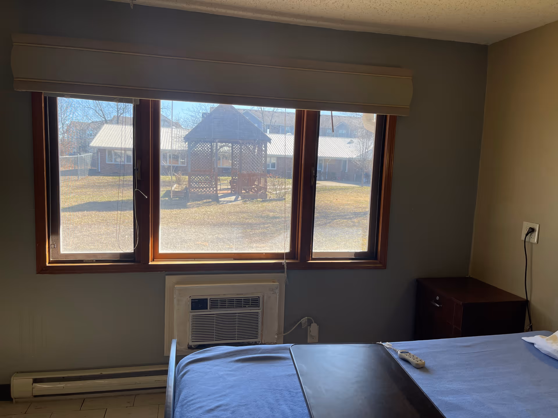 View from inside a bedroom showing a bed with a blue blanket, a bedside table, a window air conditioning unit, and a large window with a view of an outdoor gazebo and buildings in the background.