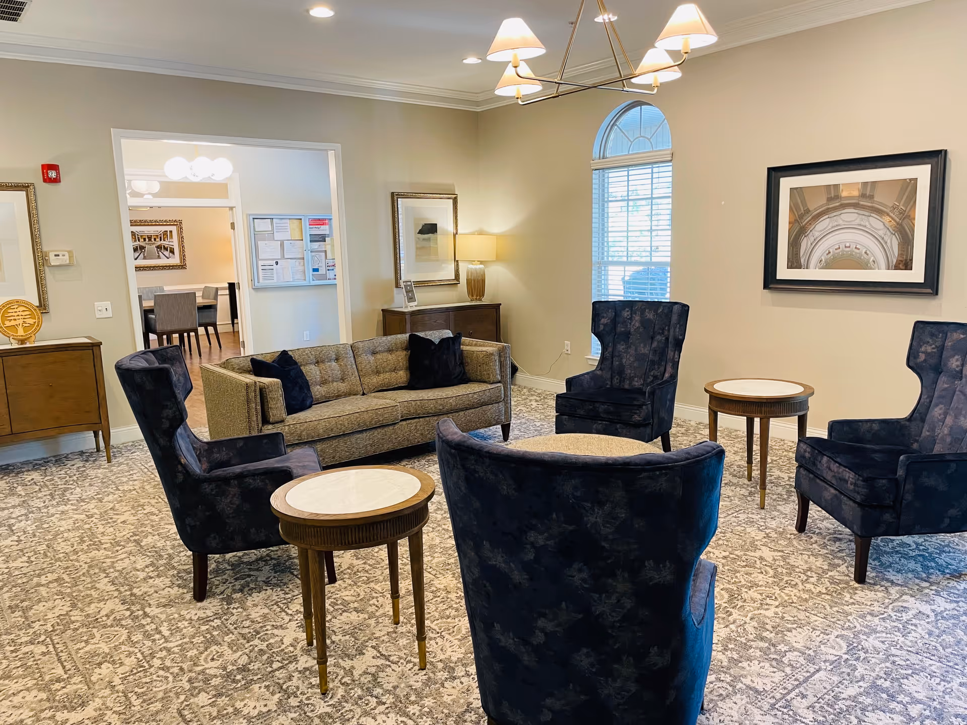 A cozy living room area with a beige sofa and four dark blue armchairs arranged around two small round tables with marble tops. The room has a patterned carpet, a large window with white blinds, a chandelier with lampshades, framed artwork on the walls, and a wooden sideboard with a lamp and decorative items. In the background, there is a view into a dining area with chairs and tables.