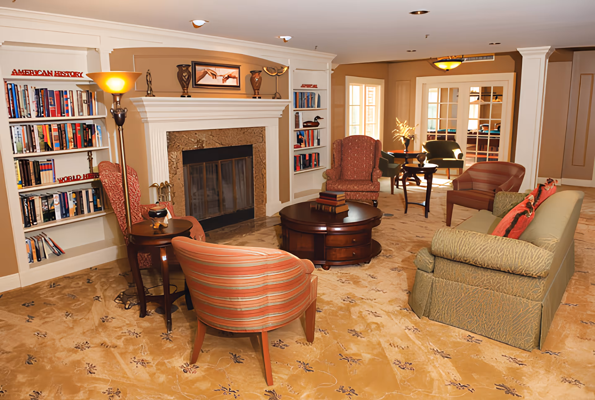 A cozy living room area in Mallard Cove featuring a fireplace with a mantle decorated with vases and a framed picture. Surrounding the fireplace are built-in bookshelves filled with books. The room has multiple upholstered chairs and a sofa arranged around a round wooden coffee table with books on it. The carpet is beige with a subtle floral pattern, and there is a floor lamp providing warm lighting. In the background, there are glass-paneled double doors leading to another room with additional seating.