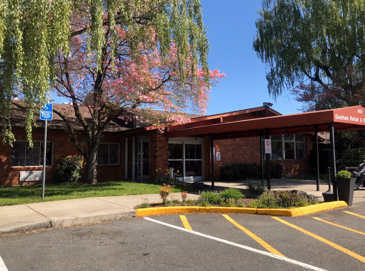 Brick single-story rehab facility entrance with a red covered canopy, flowering trees, and a handicap parking area.