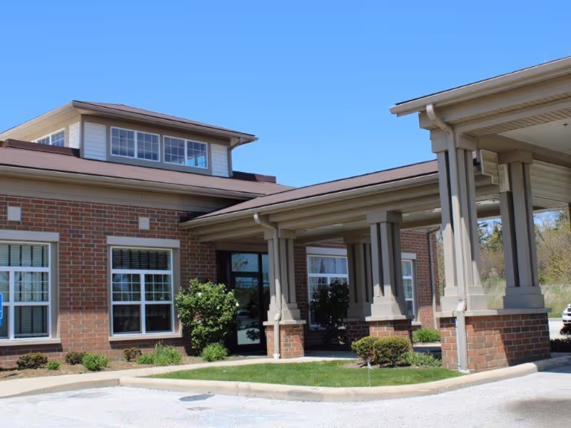Front entrance of a brick care facility featuring a covered porte-cochere, windows, and landscaping under a clear blue sky.